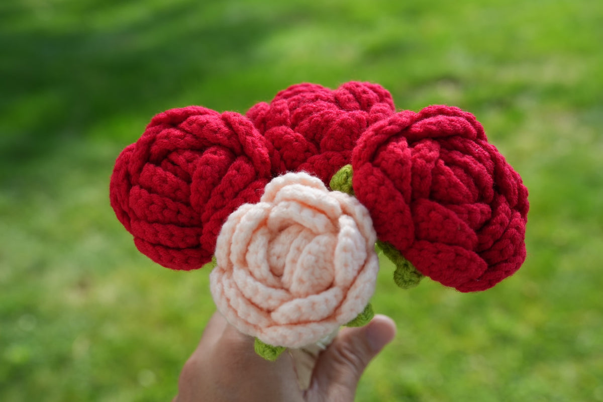 A hand holding red and pale peach crochet roses against a soft green outdoor background